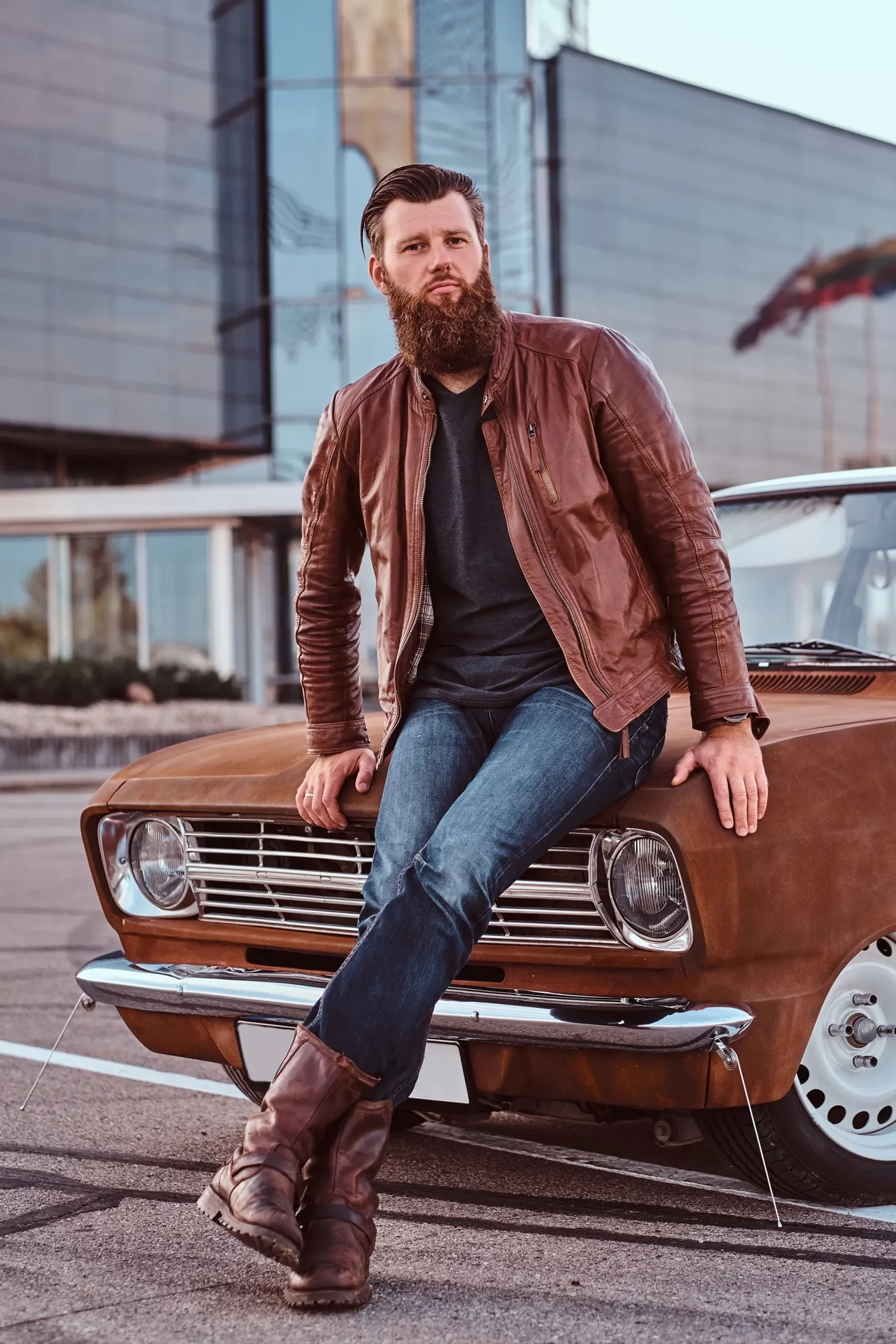 country-style-concept-bearded-male-dressed-in-brown-leather-jacket-and-boots-leaning-on-tuned-retro-car-in-the-city-parking-near-skyscraper